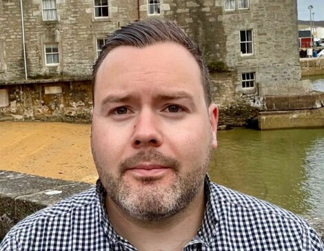 A man with short hair and a trimmed beard wearing a black and white checkered shirt stands outdoors near a stone building by the water.