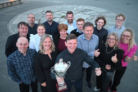 A group of people standing outdoors, smiling and giving thumbs up, with one person holding a large trophy at the front.