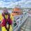 A man in a yellow waterproof suit and red life jacket stands on a dock beside an orange lifeboat on a sunny day.
