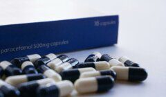 A close-up of black and white paracetamol 500mg capsules scattered in front of a blue box labeled "16 capsules.