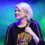 A woman with short blonde hair holds a violin on stage, wearing a black top, against a blue and green lit background.