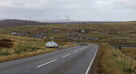 A white car is parked on the side of a rural road with scattered houses and wind turbines visible on rolling hills in the background.
