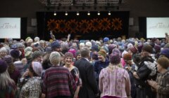 A large group of people wearing patterned wool hats and sweaters gather indoors under stage lights, facing various directions and engaging with each other.