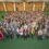 A large group of people poses together indoors on a green floor, facing the camera and smiling. The setting appears to be a spacious hall.