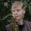 Person with short blonde hair holding a brass saxophone stands among green leaves, looking slightly to the side.