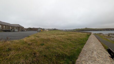 A grassy area bordered by a stone wall runs alongside a paved parking lot on the left and a coastal path on the right, with cloudy skies overhead.