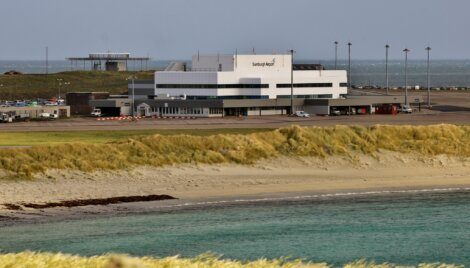 A small airport terminal building labeled "Sumburgh Airport" stands near a beach with grassy dunes and clear blue water in the foreground.