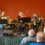 Five musicians perform on stage with accordions, a violin, and a keyboard in front of an audience, against a plain orange wall.