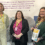 Three women stand in front of a display about Shetland’s industrial surplus, one holding a report titled “Shetland's Future.” All wear conference badges and business attire.
