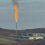 A large industrial gas flare burns with black smoke rising above a facility on a grassy, hilly landscape under a cloudy sky.