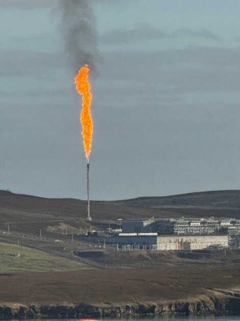 A large industrial gas flare burns with black smoke rising above a facility on a grassy, hilly landscape under a cloudy sky.