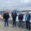 Four people stand on a dock in front of several docked boats and ships under a cloudy sky. Industrial buildings are visible in the distance.