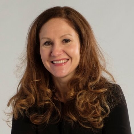 A woman with long, curly brown hair wearing a black top is smiling at the camera against a plain light background.