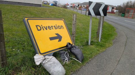 A yellow "Diversion" road sign with a right arrow is propped up on grass with sandbags, next to a road curve sign by a roadside fence.