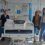 Four women stand around a hospital bed in a brightly lit hospital room, with medical equipment and chairs visible in the background.