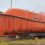 An orange enclosed lifeboat is placed on supports on grassy ground, with rescue equipment and a netted buoy nearby.