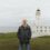 An older man stands outdoors with his hands in his jacket pockets; a white lighthouse and building are visible in the background near the sea.
