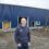 A man wearing glasses and a hoodie stands in front of a blue industrial building under construction, with construction equipment and materials visible nearby.