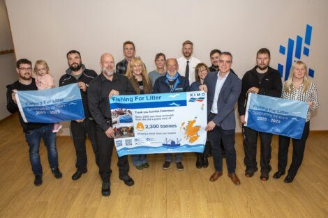 A group of people stands indoors holding a large "Fishing For Litter" banner, celebrating 20 years and 23,000 tonnes of marine litter removed from Scottish seas.