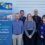 Six people stand in front of a light-colored wall next to a sign for the Brae Rural Energy Hub, which promotes climate-conscious community initiatives in Shetland.