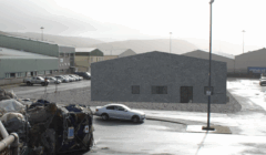 A gray, windowless building stands in an industrial parking lot with a few parked cars and stacked materials in the foreground.