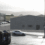 A gray, windowless building stands in an industrial parking lot with a few parked cars and stacked materials in the foreground.