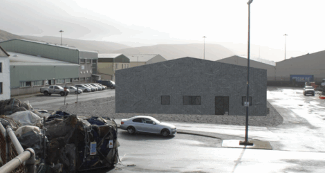 A gray, windowless building stands in an industrial parking lot with a few parked cars and stacked materials in the foreground.