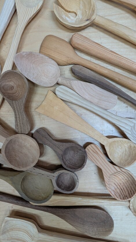 A variety of unfinished wooden spoons and utensils are scattered on a light wooden surface.