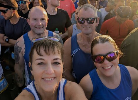 Group of runners in athletic gear pose for a selfie at a race event, surrounded by other participants, in early morning sunlight.