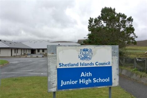 A sign for Aith Junior High School, part of Shetland Islands Council, stands in front of the school building on a cloudy day.
