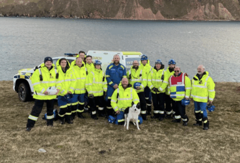 A group of rescue workers in yellow jackets and helmets stand on grass by a body of water, with a dog and emergency vehicles visible behind them.
