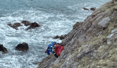 A rescue worker in blue gear assists a person secured in a red stretcher on a steep rocky cliff above the ocean.