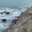 A rescue worker in blue gear assists a person secured in a red stretcher on a steep rocky cliff above the ocean.