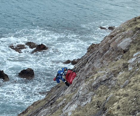 A rescue worker in blue gear assists a person secured in a red stretcher on a steep rocky cliff above the ocean.