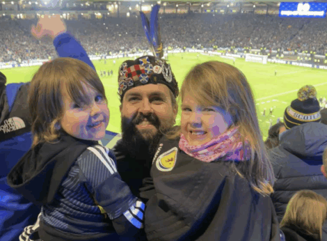 A man wearing a decorated tam o’ shanter and two children smile at the camera in a crowded football stadium.
