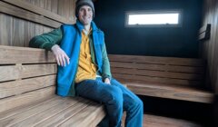 A person sits on a wooden bench inside a modern, wooden-paneled sauna with a small window letting in light.