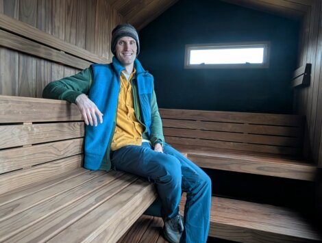 A person sits on a wooden bench inside a modern, wooden-paneled sauna with a small window letting in light.