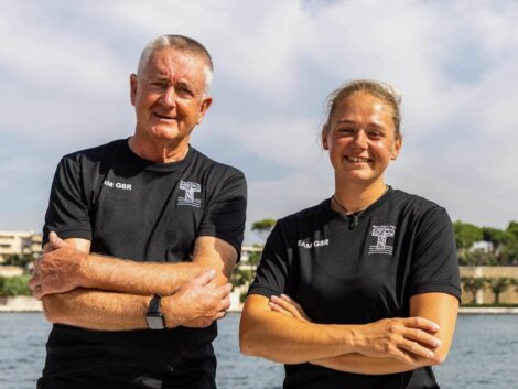 Two people in black team shirts stand side by side with arms crossed, smiling outdoors near water on a sunny day.