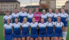 A women's field hockey team poses together on an outdoor turf field, wearing matching blue and white uniforms; one player stands in goalkeeper gear.