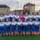 A women's field hockey team poses together on an outdoor turf field, wearing matching blue and white uniforms; one player stands in goalkeeper gear.
