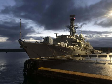 A large naval warship is docked at a pier during dusk, with cloudy skies and water reflecting the evening light.