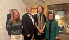 Four women stand indoors, posing for a group photo in a wood-paneled room with framed pictures and a lamp in the background.