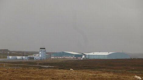 A small airport with blue buildings, including a control tower and large hangars, set in a grassy, open landscape under an overcast sky.