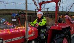 A child wearing a Santa hat and neon safety suit sits on a red Massey Ferguson tractor decorated with Christmas lights and tinsel, outdoors near a playground.