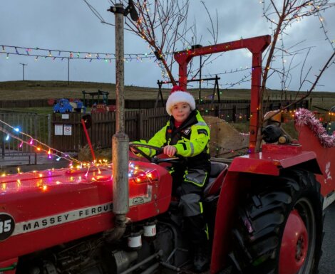A child wearing a Santa hat and neon safety suit sits on a red Massey Ferguson tractor decorated with Christmas lights and tinsel, outdoors near a playground.