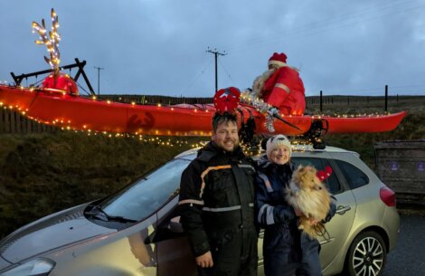 Two people and a dog stand by a car with a red kayak on its roof, decorated with lights, a Santa figure, and antlers on a cloudy day.