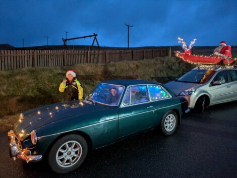Two people sit in a dark green classic car, while a person in a Santa hat stands nearby. Behind them is a silver car decorated with a Santa and sleigh display on its roof.
