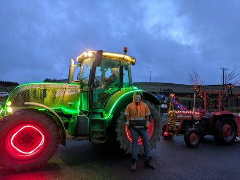 A person in a reflective jacket stands in front of a large tractor decorated with colorful lights, with a smaller, also lit-up tractor behind, at dusk.