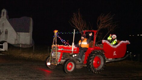 A man drives a red tractor decorated with lights, pulling a sleigh with two people in festive attire at night near a white church.
