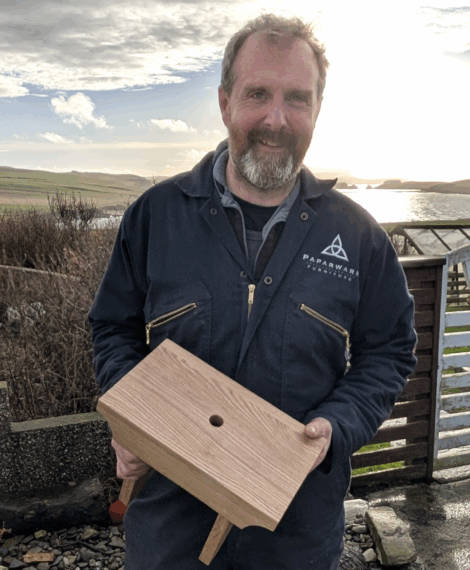 A person in a blue coverall stands outdoors, holding a wooden box with a circular hole in the center and two short legs, with fields and water in the background.